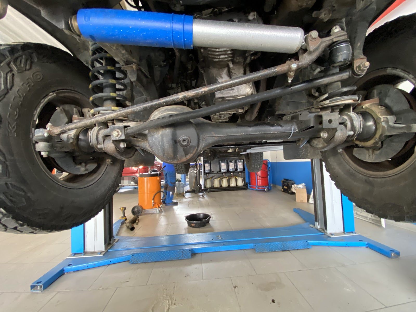 Essieu avant d'un 4x4 vu du dessous, sur un pont élévateur bleu dans l'atelier